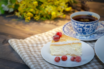 On the saucer is a piece of cheesecake cake and red berries, the whole cake on a white plate, next to it is a white tea pair with a blue pattern, everything is on a linen napkin.