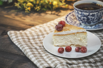 On the saucer is a piece of cheesecake cake and red berries, the whole cake on a white plate, next to it is a white tea pair with a blue pattern, everything is on a linen napkin.