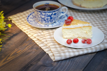 On the saucer is a piece of cheesecake cake and red berries, the whole cake on a white plate, next to it is a white tea pair with a blue pattern, everything is on a linen napkin.