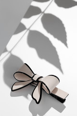 Subject shot of a cambered hair clip made of light beige glazy material in the form of a bow-knot with a black stripe. The image is made in the studio on the white surface with shadows of the leaves. © RedUmbrella&Donkey