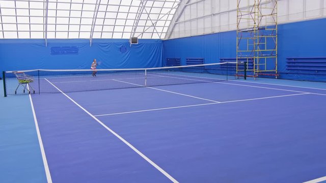 Young female tennis player training on indoor court, practicing serve and forehand