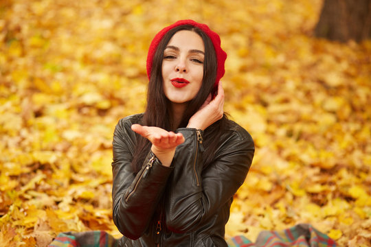 Outdoor autumn fashion image of pretty woman wearing red beret and leather jacket, sending air kiss to camera, being in motive mood, adorable female spending warm autumn day in park, bright colors.