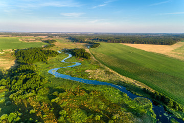 A small river flowing through meadows and agricultural fields. Aerial view. Evening shot with the setting sun.