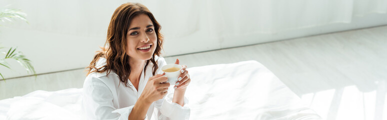 panoramic shot of attractive woman in white shirt smiling and holding cup at morning