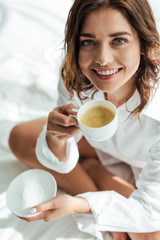 high angle view of attractive woman in white shirt smiling and holding cup at morning
