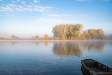 Autumn calm morning on meadow with a boat on the river. Beautiful sunrise over field with sunlight and mist.
