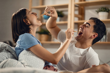 Playful couple feeding each other with a cookie in bedroom.