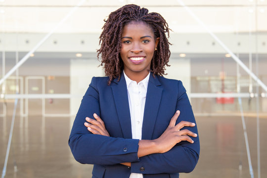 Happy Successful Business Leader Posing Near Outside. Young Business Woman With Arms Folded Standing Near Glass Wall, Looking At Camera, Smiling. African American Businesswoman Concept