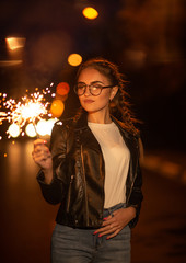 young girl with glasses stands at night on the road with sparklers in her hands