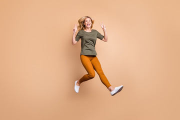 Fototapeta na wymiar Full length photo of crazy lady jumping high showing v-sign symbol saying hello friends excited summer mood wear green t-shirt yellow trousers isolated beige color background