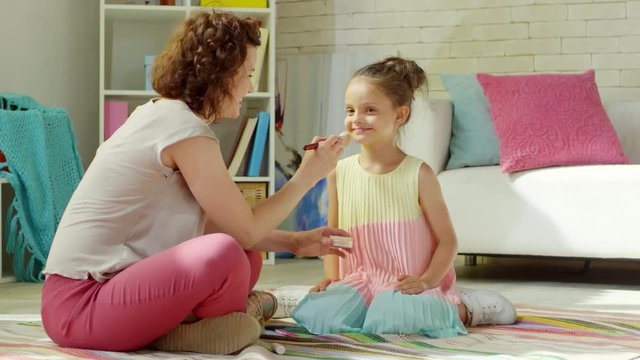 Cheerful mother applying powder to face of adorable little daughter and then tickling her with brush while playing on the floor in kids room