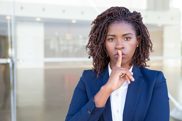 Fototapeta na wymiar Serious silent professional keeping secret. Young black business woman standing at outdoor glass wall, applying index finger at mouth, making shh gesture. Conspiracy concept