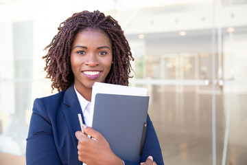 Happy friendly office assistant posing outside. Young black business woman standing at glass wall, holding documents, looking at camera, smiling. Assistant or paperwork concept