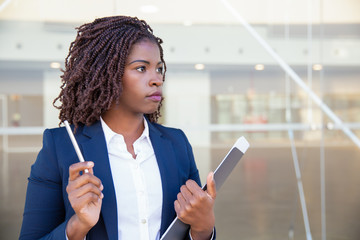 Pensive serious manager with documents waiting partners outside. Young black business woman standing at outdoor glass wall, holding pen and papers, looking into distance. Paperwork concept