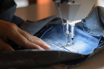 Hand of the seamstress is using a white industrial sewing machine to sew blue jeans.
