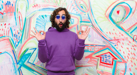 young bearded crazy man feeling shocked, amazed and surprised, showing approval making okay sign with both hands against graffiti wall