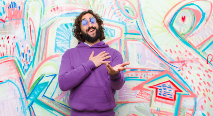 young bearded crazy man feeling happy and in love, smiling with one hand next to heart and the other stretched up front against graffiti wall