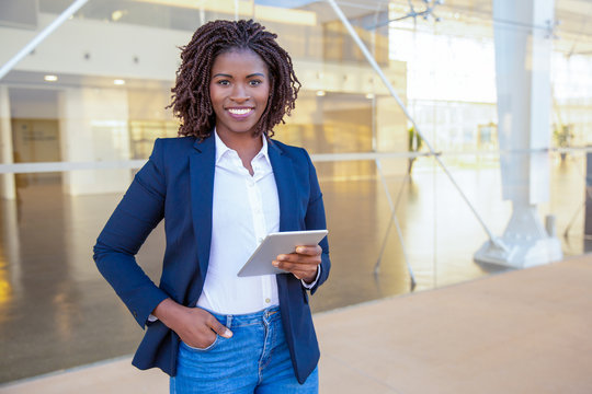 Happy professional using tablet near office building. Young African American business woman standing outside, holding digital device, looking at camera, smiling. Wi-Fi concept