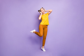 Fototapeta na wymiar Full size photo of joyful teenager lady listening modern youngster playlist music through cool earflaps wear yellow t-shirt trousers isolated pastel purple background