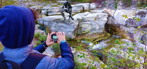 Little boy photographing a robot, on a rock, near a small river.