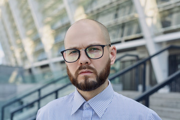 A pensive business man with brown beard stands in glasses on the street on the steps against the background of a building in the city. Business man, employee, programming. Studying.