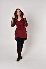 full length portrait of a brunette girl wearing a red fantasy tunic with hood. Standing pose on a white studio background