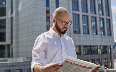 A horizontal shot of a bald young guy in glasses with a beard in a white shirt is standing with a newspaper in his hands and carefully reads it against background of surrender in the city. Busy.