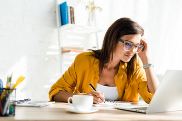 attractive female freelancer working on laptop and writing in home office