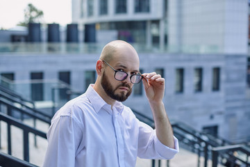 A bald guy holding the glasses with a dark beard on the street on the steps against the background of a building in the city. Worker, employee, employment