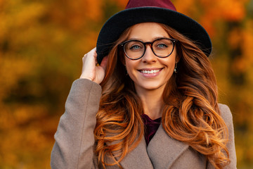 Portrait of a beautiful happy young woman with a lovely smile with an elegant hat in stylish glasses in a fashionable coat in the park.Cheerful hipster girl in trendy outerwear posing in autumn forest