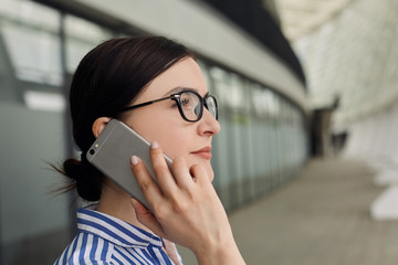 Intelligent business woman with brown hair in glasses in a blue and white striped shirt with white cuffs on her sleeves calling on gray cell phone against the background of the city. Busy.
