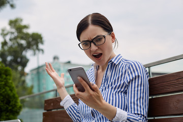 A pretty business woman with dark tied hair in glasses and a striped shirt sits in a park on a bench and looks in surprise at the gray phone in hand. Background of the city.