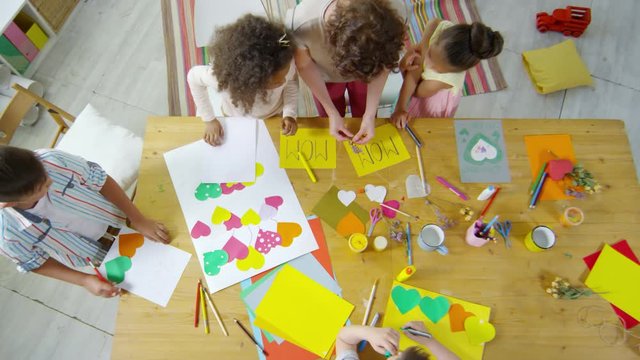 Top view zoom out shot of multi-ethnic group of little kids making colorful greeting cards for their mothers with help of female teacher in kindergarten