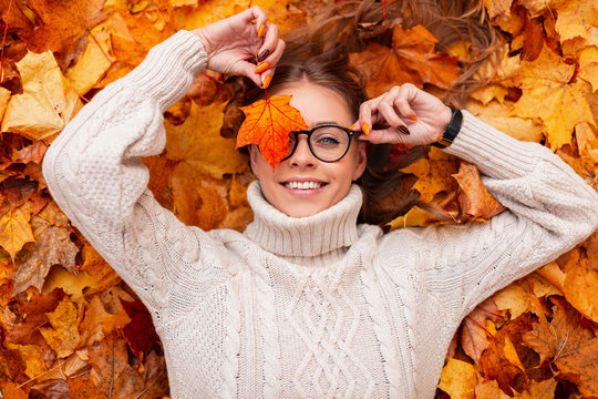 Funny young hipster woman with a positive smile with an orange maple leaf in glasses in a trendy knitted sweater lies on autumn foliage in the park. Joyful girl holds in a hand golden leaf near face.