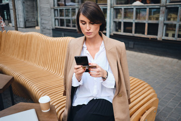 Young confident stylish businesswoman with credit card using cellphone during coffee break in cafe...