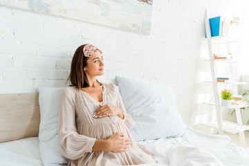 pregnant woman in sleeping mask touching her belly while lying in bed in the morning