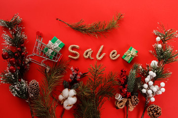 Christmas sale typography on the red background among fir tree branches, shopping cart, gift boxes. Advertising concept.