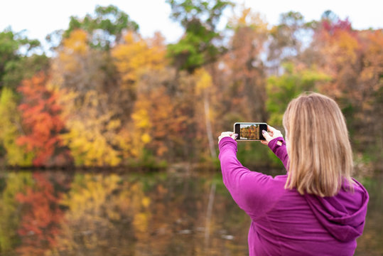 woman taking photo of fall colors along lake with smartphone