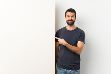 Young handsome man with beard holding a big empty placard pointing to the side to present a product