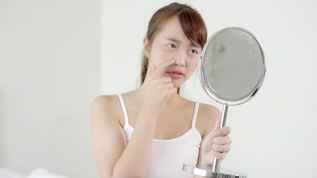 Beautiful young asian woman looking the mirror and touch face is a acne, zit treatment, girl problem beauty and skin, perfect with wellness in the bedroom at home with skin care and health concept.