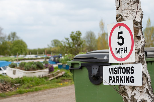 Close-up if a 5mph speed limit seen seen at the entrance to a boat yard and marina. Boats can be seen moored across the road, into the distance.