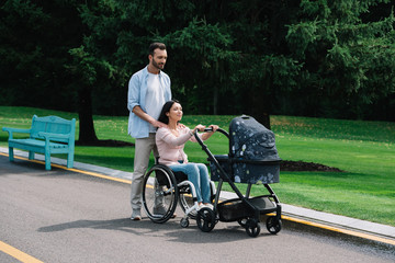 happy woman in wheelchair walking with husband and baby carriage in park