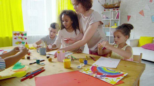 Group of multi-ethnic little children closing gouache paints while helping female teacher with cleaning table after drawing lesson in art class