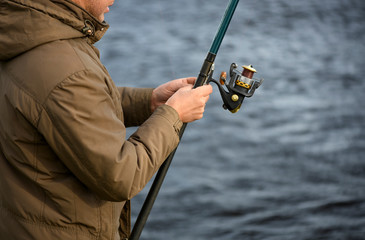 The caucasian fisherman is standing near a water and holding the fishing rod in his hands in a cool windy day.