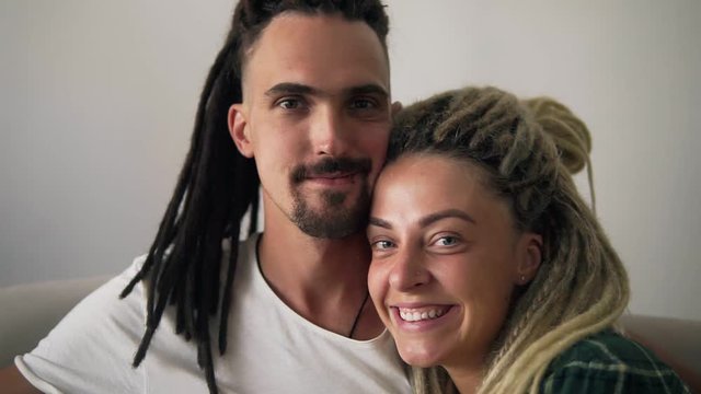 Close up portrait of the young dreadlock couple in love looking directly to the camera and smiling on the white background wall. Indoors