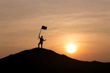 Silhouette of young man holding a flag on top mountain, sky and sun light background. Business success and goal concept.