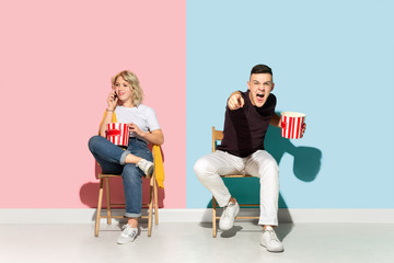 Young emotional man and woman in bright casual clothes posing on pink and blue background. Concept of human emotions, facial expession, relations, ad. Beautiful couple watching cinema with popcorn.