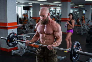 Naklejka na ściany i meble Bald man with a beard in the gym. Muscular bodybuilder guy doing barbell exercises. Strong man with a naked torso. The young athlete is preparing for weightlifting competitions.