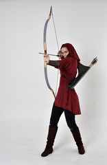 full length portrait of a brunette girl wearing a red fantasy tunic with hood, holding a bow and...