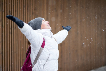 Young smiling caucasian girl dressed in white jacket enjoying first snow with closed eyes and rised hands